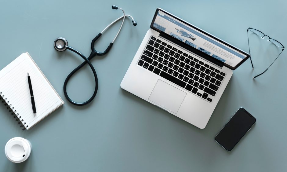 From left to right, a notebook and pen, a stethoscope, laptop, phone and pair of glasses on a table.