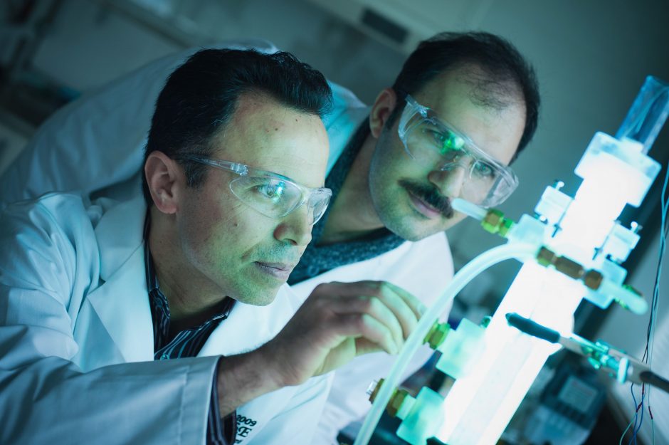 Two researchers wearing lab coats and safety goggles examine a glowing green piece of equipment in a lab.